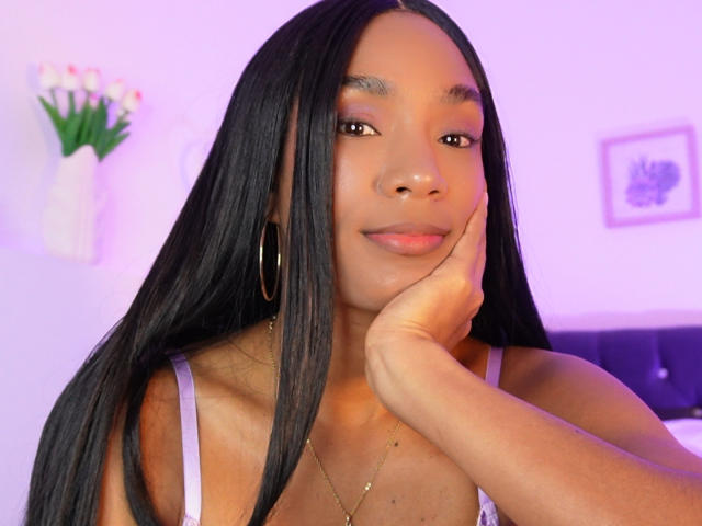NatLewis has long black hair, brown eyes, and a cute smile, resting her chin on her hand, wearing a white tank top, and gold hoop earrings. Her tits are small.