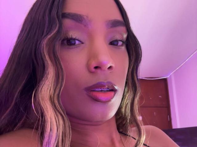 NatLewis has wavy brown hair with blonde highlights, purple lipstick, and brown eyes, looking at the camera with a slightly open mouth in a purple-lit room.