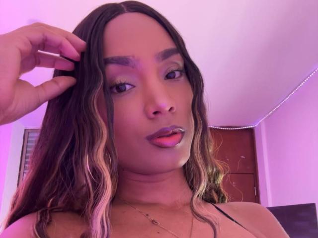 NatLewis with wavy brown hair, purple eyeshadow, and pink lipstick, tongue slightly out, hand in hair, wearing a black spaghetti strap top, in a pink-lit room.