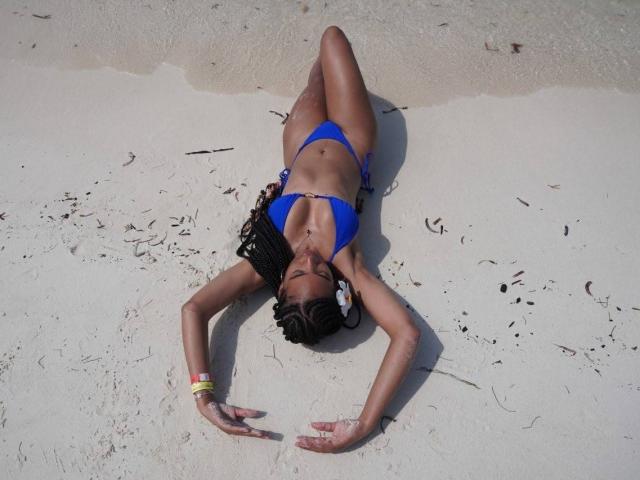 NatLewis with dark skin, blue bikini, and braided hair lies on sandy beach, arms stretched above head, legs slightly apart, wearing a yellow bracelet, face down.