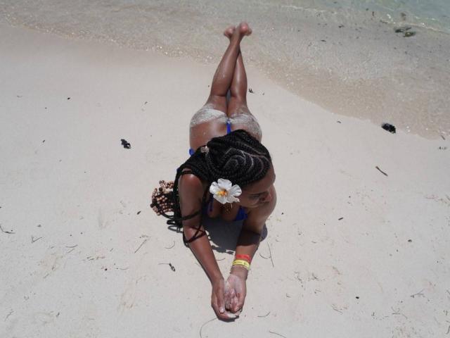 NatLewis, dark-skinned, lying on her back on the beach, legs up, wearing a blue bikini, white flower in her braided hair, lace panties, red and yellow bracelet, hands on sand.