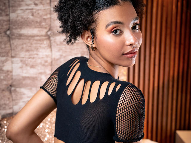 Kiara Kamau mit lockigem, schwarzem Haar, brauner Haut, trägt ein schwarzes, netzförmiges Oberteil mit Ausschnitten, das ihren Rücken zeigt und sie blickt über ihre Schulter, lächelnd. Ihre kleinen Brüste sind sichtbar..