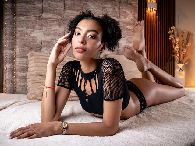 KiaraKamau lying on a bed, wearing a black fishnet top with cutouts and black panties, her curly hair, brown skin, and gold watch, legs up, looking at camera.
