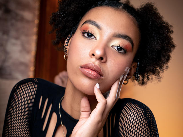 Kiara Kamau mit lockigem, schwarzem Haar, die nach unten schaut, trägt ein schwarzes Netzoberteil, goldene Ohrringe, weißen Nagellack und orangefarbenen Lidschatten. Ihre Hand berührt ihre Wange, ihre Haut ist weich und ihre Lippen sind leicht geschürzt..