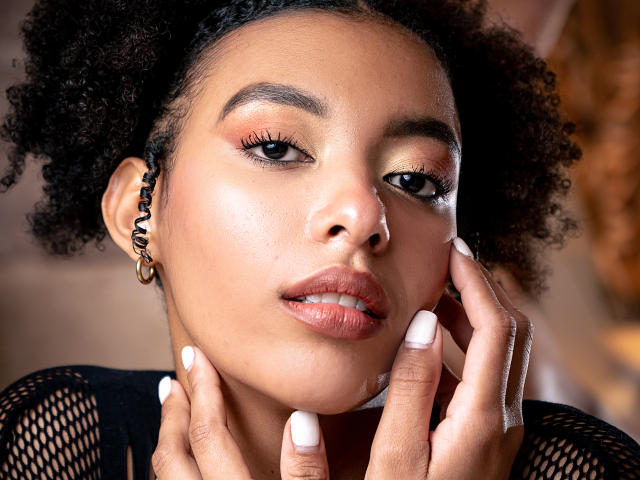 KiaraKamau's face, dark curly hair, brown skin, pink eyeshadow, white nails, slightly parted glossy pink lips, gold earring, black fishnet top, hands touching her face.
