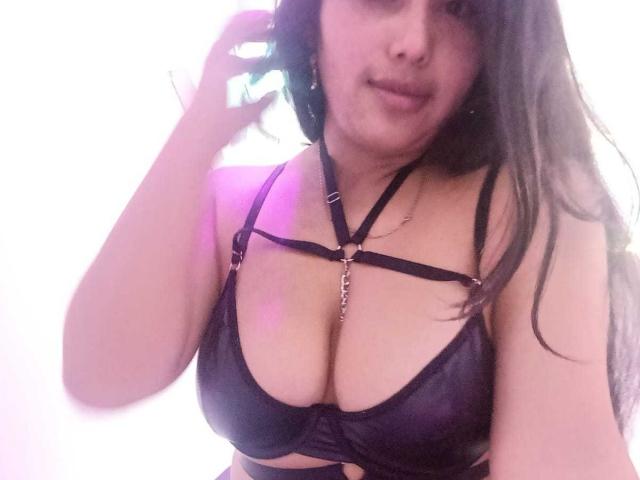 GemmaBellini with long black hair, wearing a black leather harness bra, showing her big tits, holding her hair, smiling slightly. Soft pink light on her right.