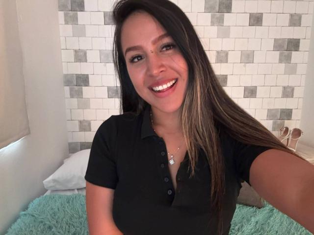 LiaDiane with long brown hair, smiling, wearing a black polo shirt, showing her tits slightly, standing in front of a white and gray tiled wall, on a bed.