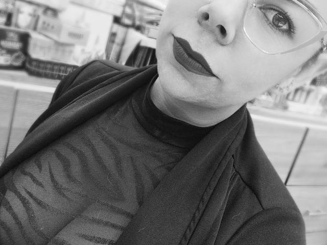 EmiliaMilf with dark lipstick, glasses, and a sheer top showing her tits, wearing a black jacket, in a black-and-white street photo.