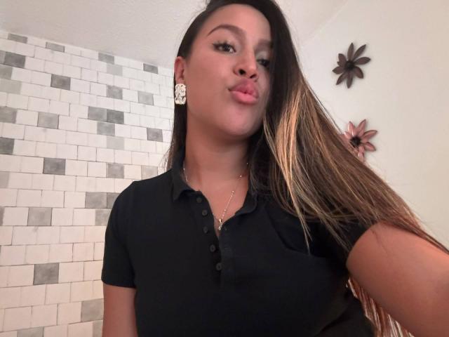 LiaDiane met volgeplooide lippen, een zwart poloshirt, lang bruin haar, witte bloemoorringen, een gouden ketting, bruine ogen, lichte huid, licht gekanteld hoofd, haar vasthoudend..