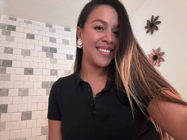 LiaDiane has long brown hair, wearing a black polo, smiling with white earrings, holding her hair, showing her tits, in a bathroom with white and gray tile.