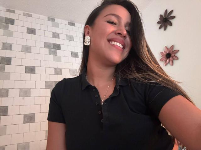 LiaDiane with long brown hair, smiling, wearing a black polo, white flower earrings, and a gold necklace, showing her cute face and tits in a selfie.