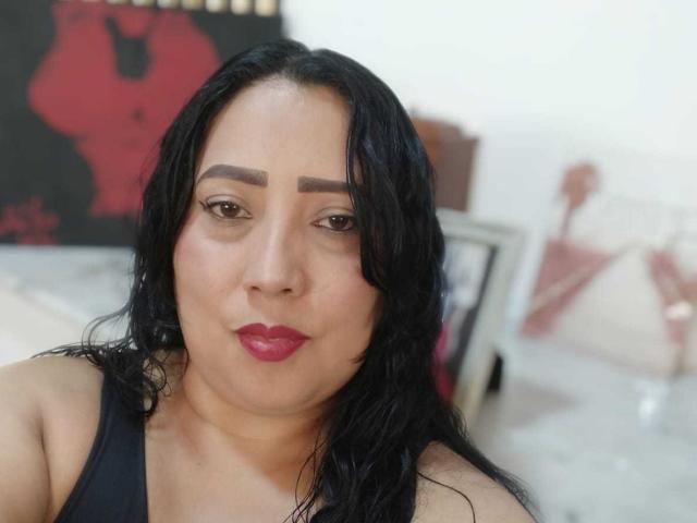 Amber Moretti mit schwarzen, welligen Haaren, brauen Augenbrauen, rotem Lippenstift und einem schwarzen Tanktop, zeigt einen ernsten Gesichtsausdruck, weiche Haut und ein leicht rundliches Gesicht..