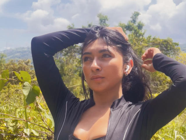 AmeliRoux has tan skin, dark hair, and is wearing a black zip-up top with arms raised, showing small tits, and wearing earbuds. She's outdoors with greenery.
