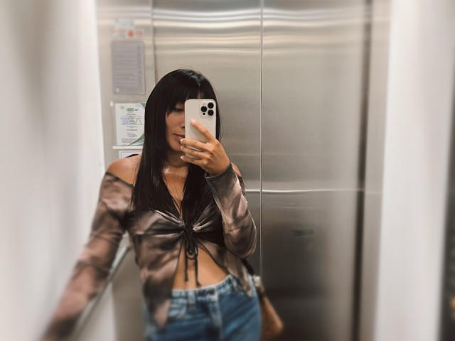 AmeliRoux with long black hair, tied-up brown and black shirt revealing her tits, and blue jeans, taking a mirror selfie in a metallic elevator, holding an iPhone.