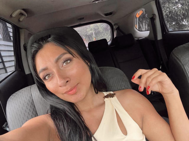 AmeliRoux with long black hair, brown eyes, and red nails, wearing a white top with cutout, sitting in a car, holding her hair, smiling slightly, showing her tits.