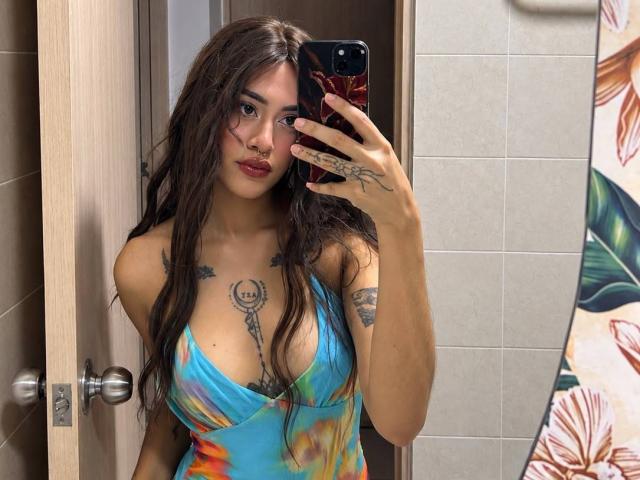 Alepeach with long brown hair, tattoos on chest and arm, wearing a blue and orange tie-dye dress, holding a phone with a red case, showing her tits.