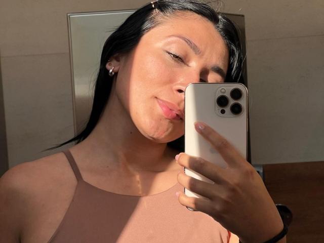 AmeliRoux with black hair, pink lipstick, and brown tank top, pouting lips, holding white iPhone, sunlight on face, pink earrings, wood floor, and subtle smile.