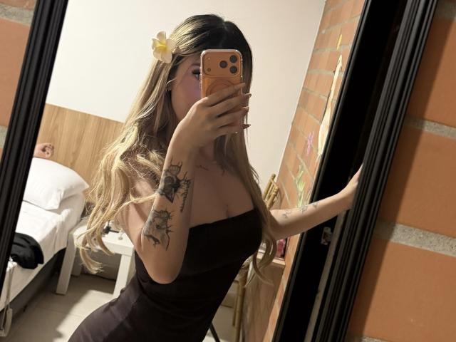 SammyViolet, con lunghi capelli biondi, un fiore giallo, un abito nero senza spalline, tatuaggi sul braccio destro, che si scatta un selfie allo specchio, mostrando il seno, tenendo in mano un telefono arancione, in una stanza d'albergo..