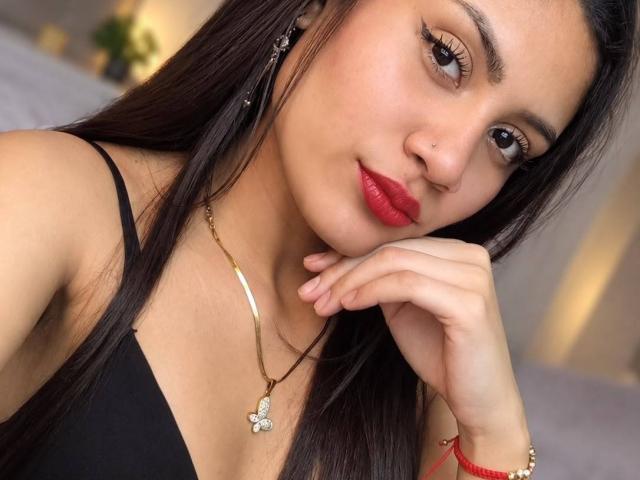 ValeryPoleth has long black hair, red lipstick, and a butterfly necklace, with her hand on her chin, wearing a black top, and a red bracelet on her wrist.