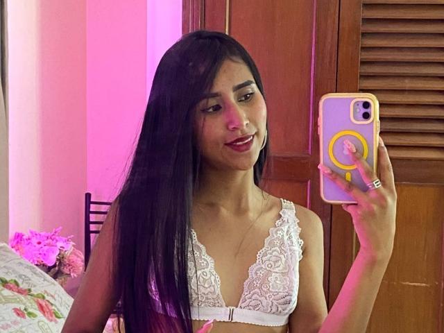 Valentina, mit langen, schwarzen Haaren, lächelnd, trägt einen weißen Spitzen-BH und hält ein Telefon mit einem gelben Ring. Sie macht ein Selfie vor einem Spiegel, auf dem ihre Brüste zu sehen sind..