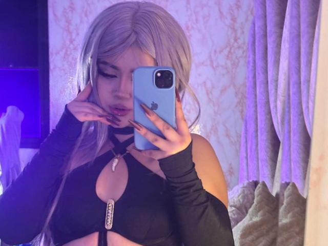 Lyanekkoo with long silver hair, black cutout top showing her tits, holding a blue phone, covering her mouth with one hand, wearing black arm sleeves, in a mirror selfie.