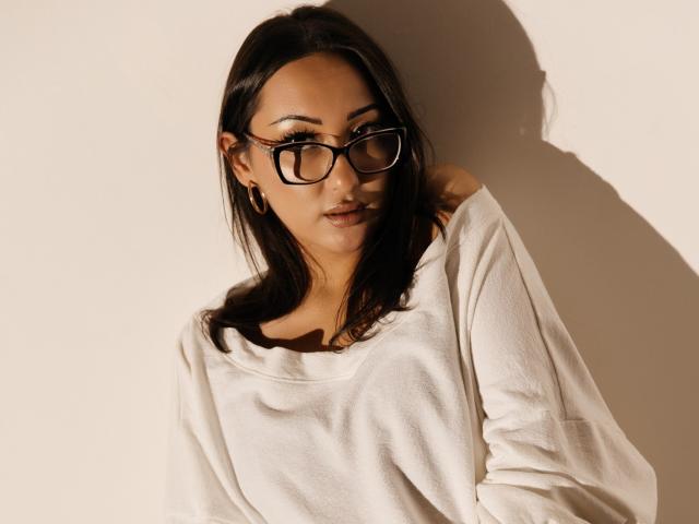 EvaSarazardje with black glasses, brown skin, and long dark hair, wearing a loose white shirt, looking slightly to the side, shadow on wall. Boobs not visible.
