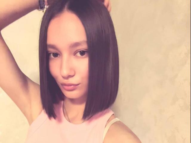 ShanGrines a une coupe de cheveux courte et noire, une peau claire et de petites seins, elle porte un haut rose sans manches, avec une expression détendue, légèrement satisfaite, et les bras levés..
