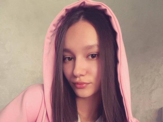 ShanGrines with long brown hair, pink hoodie, and soft brown eyes, has a cute face with slightly pouty lips, wearing the hood up, looking at the camera.