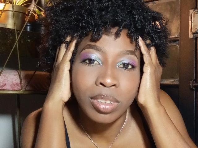 XMeryVegaXi tiene el cabello oscuro y rizado, sombra de ojos morada y azul, labios rosados brillantes y las manos en la cara. Lleva una blusa negra y un collar de plata..