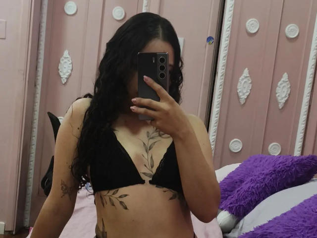Miia Bennet con reggiseno nero, capelli neri ondulati, tatuaggi sul petto e sulle braccia, che tiene un telefono, coprendosi il viso, in piedi in una stanza rosa con un cuscino viola, parete rosa..