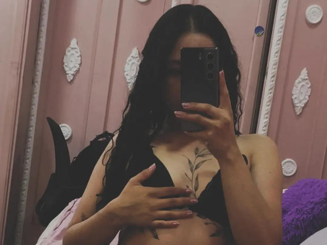 MiiaBennet met lang zwart haar, die een zwarte beha draagt, met een telefoon voor haar gezicht, waarbij ze haar borsten met één hand bedekt, en een bloemen-tatoeage op haar borst laat zien..