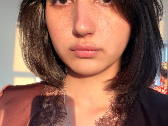 HeikeCarchi has freckled skin, a black bob, and a small beauty mark on her upper lip. She's wearing a lace-trimmed top with a reddish-brown and black combo.