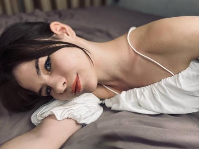 SwetHunny with dark hair, lying on a bed, wearing a white off-shoulder top, looking at the camera with a slight smile, showing her bare shoulders and subtle makeup.