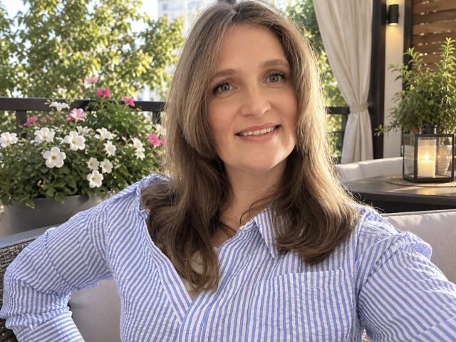 LoraParkerHot with shoulder-length brown hair, blue eyes, and a smile, wearing a blue and white striped shirt, sitting outdoors with flowers and lanterns in the background.