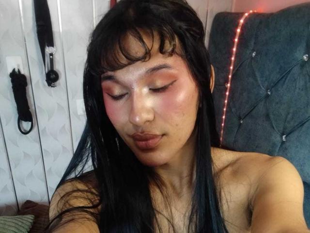 PamelaXFox with pink eyeshadow, glossy lips, and black hair, pouting with a slightly flushed face, sitting on a gray tufted headboard. Her bare shoulders are visible.