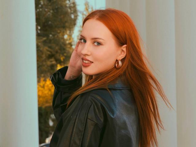 Janna Corsaro mit langen, roten Haaren, lächelnd, trägt eine schwarze Lederjacke, goldene Reifenohrringe und einen Nasenring, sie berührt ihre Haare und schaut über ihre Schulter..