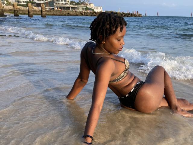 SexyArya, con piel oscura y pequeños senos, sentada en agua poco profunda, vistiendo un top de bikini de camuflaje y pantalones negros, mirando hacia un lado, con el pelo mojado y una pulsera..