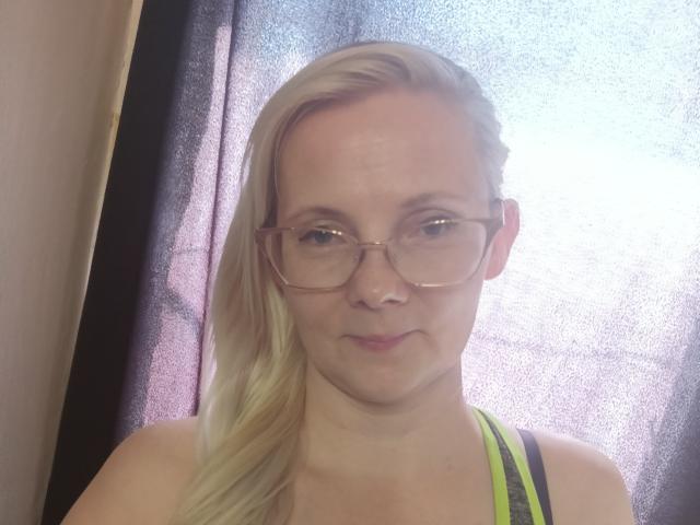 Blonde Sweetie69g with glasses, medium tits, and a subtle smile, wearing a neon green sports bra, standing in front of a sunlit window.