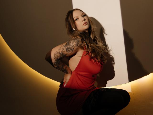 MariaRobinson with tattoos on her arm, wearing a red dress, squatting with hands on hips, black pants, brown hair, looking at camera, soft light, shadow on wall.