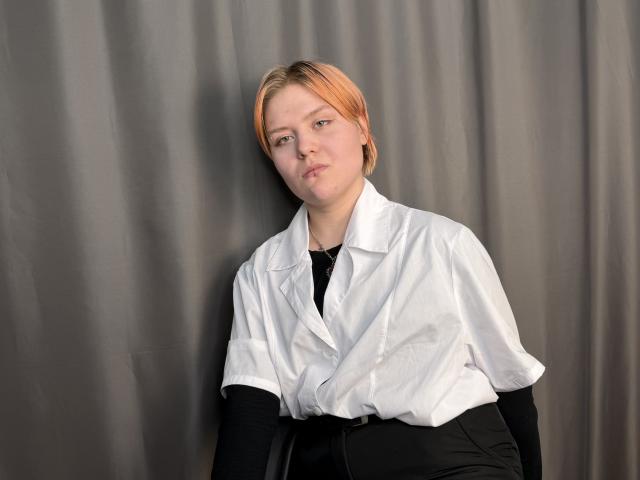 BlytheErby with light orange hair, wearing a white button-up shirt and black top, looking slightly annoyed, standing against a gray curtain. She's got a chubby ass.