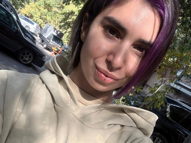 TameikaMacclellan has purple-dyed hair, a nose ring, and a lip piercing, wearing a beige hoodie, smiling with light brown eyes, and a slight tan.