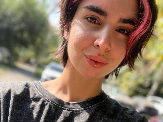 TameikaMacclellan with short brown hair, red highlights, nose ring, lip piercing, and black shirt, smiling at the camera, outdoors in sunlight.