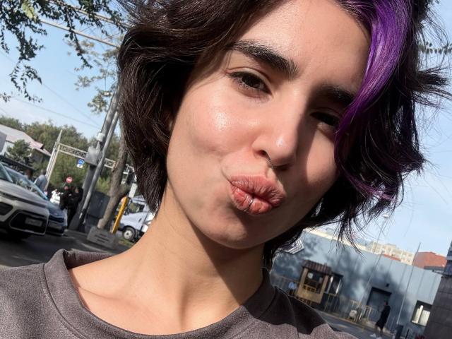 TameikaMacclellan with pouty lips, kissing air, short wavy brown hair with purple streak, brown eyes, wearing gray shirt, sunlight highlighting her skin, urban street background.