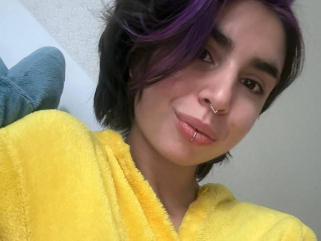 TameikaMacclellan with purple-streaked black hair, nose ring, lip piercing, and yellow fuzzy robe, smiling softly at the camera.