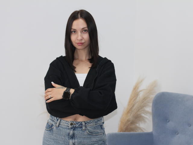 LivaLoy mit dunkelbraunen Haaren, schwarzer, kurzer Kapuzenjacke, weißem Oberteil, blauen Jeans, verschränkten Armen, einer schwarzen Armbanduhr, stehend, mit einem schelmischen Lächeln, ein flauschiger blauer Stuhl und getrocknetes Pampasgras im Hintergrund..