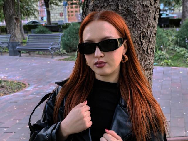 Janna Corsaro mit langen, roten Haaren, schwarzen Sonnenbrillen, einem schwarzen Rollkragenpullover, einer schwarzen Lederjacke, die sie in der Hand hält, vor einem Baum stehend, mit rosa Nagellack, kleinen Reifenohrringen, wirkt ernst..