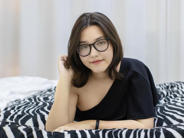 LilySinclair, mit Brille, schwarzem Hemd und mittlerer Brustgröße, liegt auf einem bett mit Zebra-Muster, die Hand im braunen Haar, und schaut in die Kamera, mit einem sanften Lächeln, vor weißem Hintergrund..