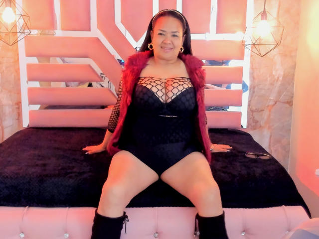 MiaaVelvett, curvy with big ass, wearing black lingerie with fishnet sleeves, red fur vest, black knee boots, sitting on pink bed, smiling, dark hair, gold earrings, warm light.