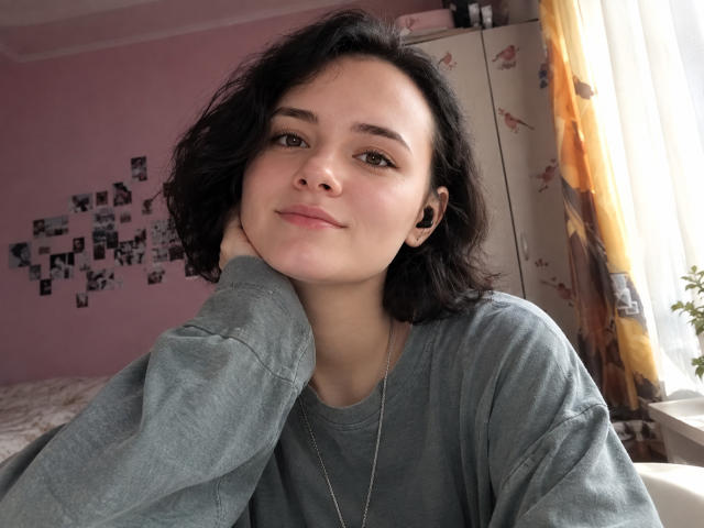 TreenaVisosky with short, curly black hair, wearing a gray sweatshirt, sitting, head on hand, small nose ring, and earbuds, smiling slightly, in a pink room.