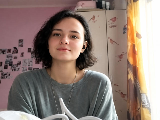 TreenaVisosky, with short black curly hair, gray sweatshirt, and necklace, sits smiling, holding a white pillow, sunlight from a yellow curtain on the right.